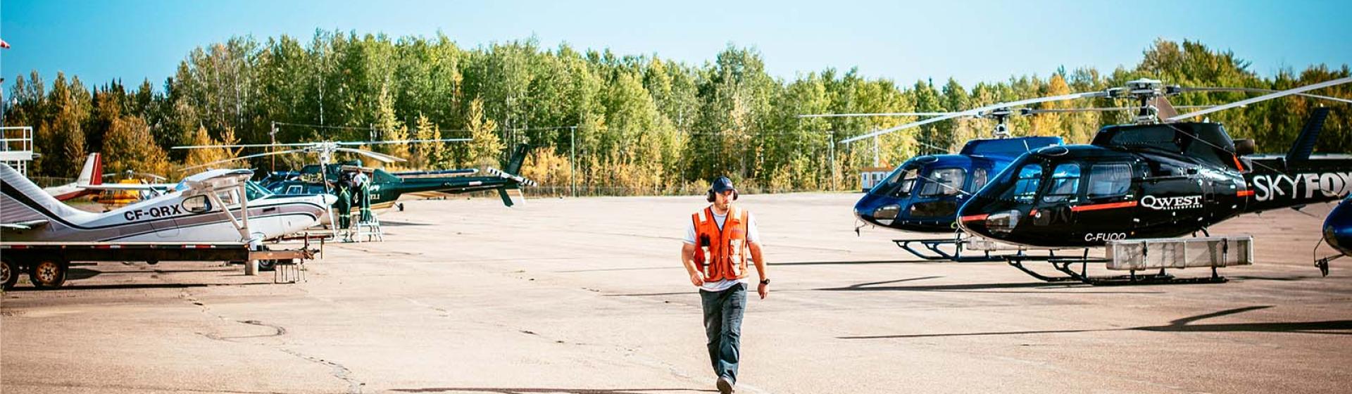 Airport worker in an orange vest walking with helicopters and planes in the background.
